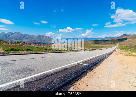 Lungo rettilineo di strada rurale che conduce verso le montagne al di là sotto cieli azzurri con soffici nuvole. Foto Stock