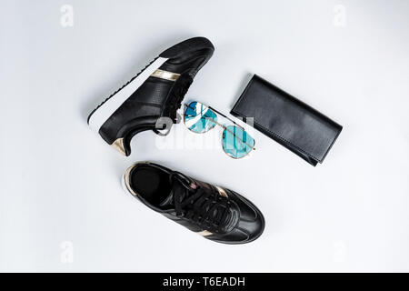 Moda sneakers nero su una suola bianca con calze nere e accenti d'oro, occhiali da sole con gli occhiali blu e un portamonete nero su sfondo bianco Foto Stock