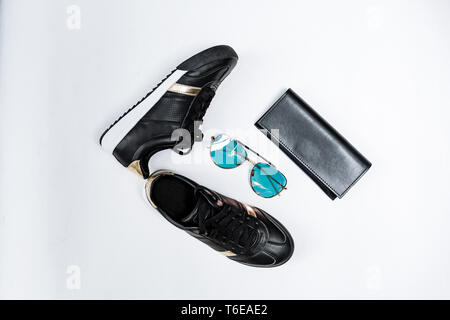 Moda sneakers nero su una suola bianca con calze nere e accenti d'oro, occhiali da sole con gli occhiali blu e un portamonete nero su sfondo bianco Foto Stock