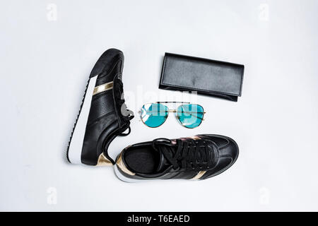 Moda sneakers nero su una suola bianca con calze nere e accenti d'oro, occhiali da sole con gli occhiali blu e un portamonete nero su sfondo bianco Foto Stock