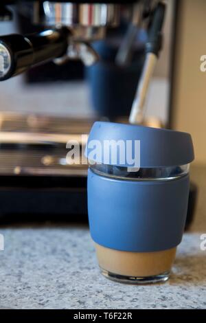 Parte anteriore vista ravvicinata di vetro da asporto tazza di caffè con manicotto in silicone e il coperchio con la macchina per espresso in background sul banco di cucina. Foto Stock