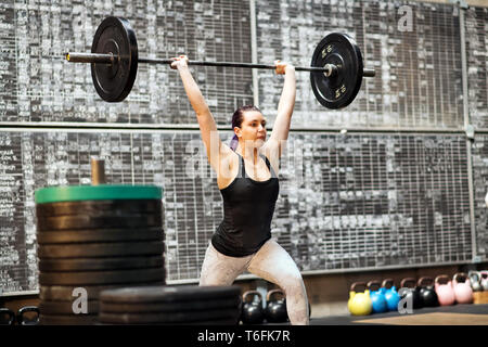 Giovane donna di eseguire una pulizia e jerk esercizio in una palestra di formazione durante il sollevamento del barbell sopra la sua testa con bracci estesi dopo il primo movimento Foto Stock