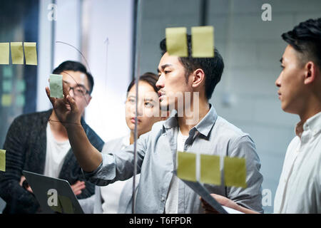 Giovani asiatici imprenditore della piccola azienda mettendo una nota adesiva sul vetro in ufficio durante la riunione del team di formulare strategie di business. Foto Stock