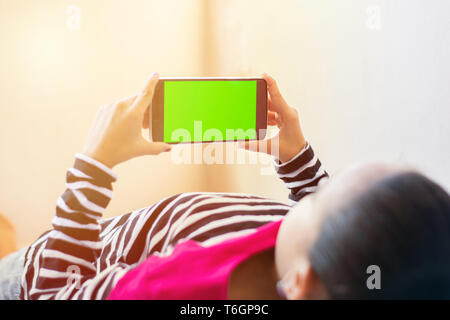 Little Indian Kid con il telefono nelle sue mani di dormire sul letto, mock-up con schermo verde, concentrarsi sul telefono Foto Stock