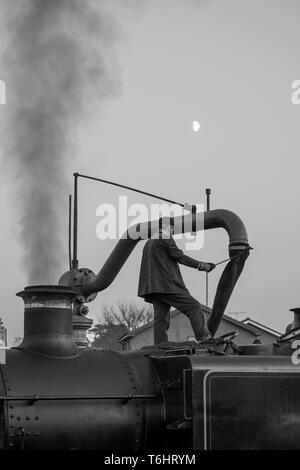 Bianco & Nero colpo di locomotiva a vapore membro di equipaggio isolato permanente sulla sommità di vintage UK motore a vapore usando acqua gru e il riempimento di un serbatoio dell'acqua. Foto Stock