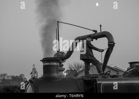 Bianco & Nero colpo di locomotiva a vapore membro di equipaggio isolato permanente sulla sommità di vintage UK motore a vapore usando acqua gru e il riempimento di un serbatoio dell'acqua. Foto Stock