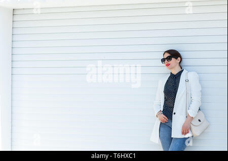 Donna elegante in occhiali da sole in piedi sul muro bianco sullo sfondo. Trendy moda casual look in copia in bianco posto nello spazio. Foto Stock