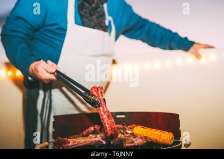 Senior la cottura della carne sul barbecue - Chef cuocere carne e salsicce che serve a cena sulla terrazza sul tetto - Concetto di cena barbecue e mangiare insieme all'aperto Foto Stock