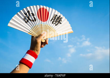Mano di sostenitore dello sport con la bandiera del Giappone di colori con la fascetta da polso mantenendo la ventola decorate con i caratteri kanji compitazione hisshō, vittoria certa in inglese Foto Stock