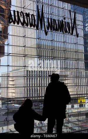 In una profila vista posteriore, un uomo che indossa un cappello da baseball mantiene la mano di una giovane ragazza come si guarda attraverso una finestra di Hudson Yards mall fuori sul temporaneamente denominata nave scultura in acciaio parzialmente visibile sulla sinistra con un alto edificio ancora in costruzione a destra e la gente nel cortile sottostante. Sopra le loro teste è il lato inverso di un Neiman Marcus department store in segno. L'Hudson Yards complesso, che ha debuttato il 15 marzo 2019, a controversie circa il fatto che esso era troppo elitario, abbraccia retail residenziale, di ristoranti e di istituzioni culturali e spazi pubblici. Foto Stock