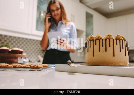 Pasticceria in cucina con lo chef pasticceria utilizzando il telefono cellulare e la tavoletta digitale. Pasticceria prendendo gli ordini sul telefono. Foto Stock