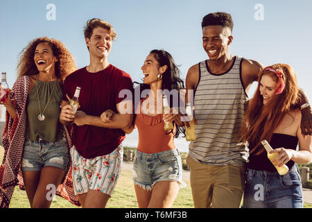 Gruppo di amici camminare insieme con bracci di bloccaggio. Giovani uomini e donne con birre camminare insieme e divertirsi. Foto Stock