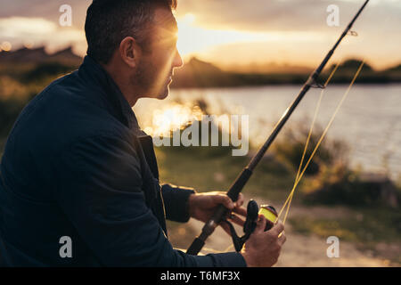 Close up di un uomo seduto vicino a un lago in possesso di una canna da pesca. Vista laterale in prossimità di un uomo la pesca nei pressi di un lago con il sorgere del sole in background. Foto Stock