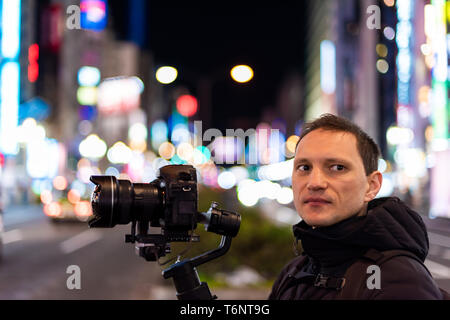 Tokyo, Giappone quartiere Shinjuku di notte con il giovane fotografo ritratto videografo riprese con la telecamera e con sospensione cardanica di sfondo bokeh di fondo paglierino Foto Stock