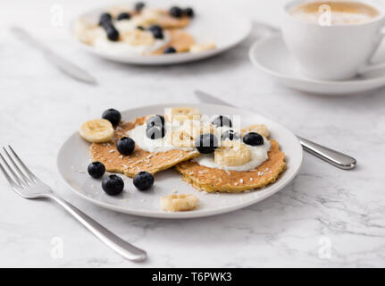 Pancake con mirtilli, banana e yogurt sulla parte superiore. Due piastre di colazione casalinga con frutti di bosco e la tazza di caffè al tavolo di marmo. Foto Stock