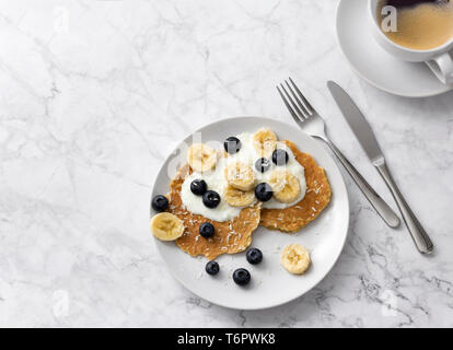 Pancake con mirtilli, banana e yogurt sulla parte superiore. Piastra della colazione casalinga con frutti di bosco e la tazza di caffè al tavolo di marmo. Foto Stock