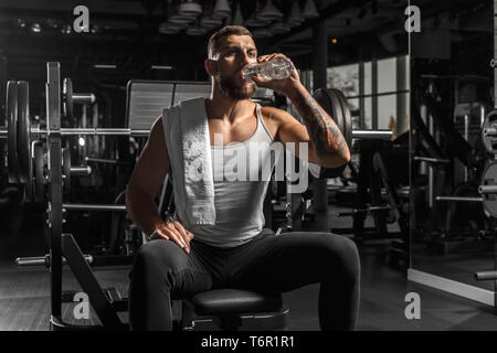 Uomo Barbuto bodybuilder seduta sul banco a palestra con asciugamano vaso di contenimento acqua potabile sete Foto Stock