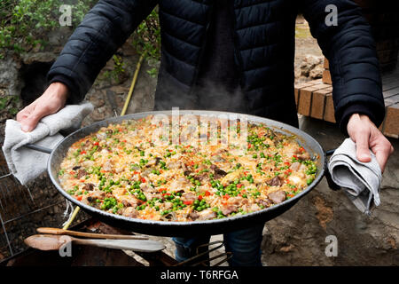 Primo piano di un giovane uomo caucasico con una tipica paella spagnola, cotto in una padella per paella con fuoco di legna, all'aperto Foto Stock