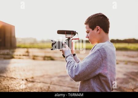 Giovane maschio videografo studiando e utilizzando la fotocamera mirrorless con microfono accessorio per la creazione di un video. Tramonto con candelette mette in evidenza il contrasto. Foto Stock