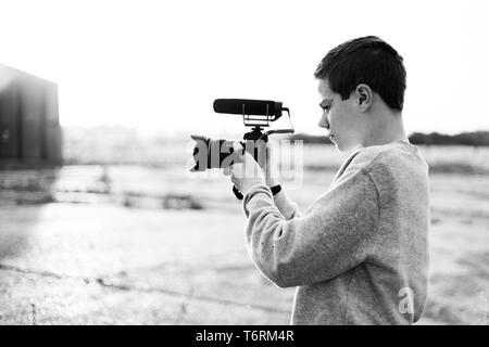 Giovane maschio videografo studiando e utilizzando la fotocamera mirrorless con microfono accessorio per la creazione di un video. Tramonto con candelette mette in evidenza il contrasto. Foto Stock