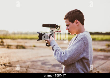 Giovane maschio videografo studiando e utilizzando la fotocamera mirrorless con microfono accessorio per la creazione di un video. Tramonto con candelette mette in evidenza il contrasto. Foto Stock