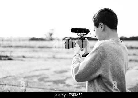 Giovane maschio videografo studiando e utilizzando la fotocamera mirrorless con microfono accessorio per la creazione di un video. Tramonto con candelette mette in evidenza il contrasto. Foto Stock