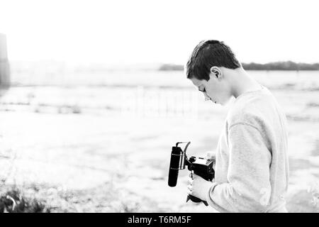 Giovane maschio videografo studiando e utilizzando la fotocamera mirrorless con microfono accessorio per la creazione di un video. Tramonto con candelette mette in evidenza il contrasto. Foto Stock