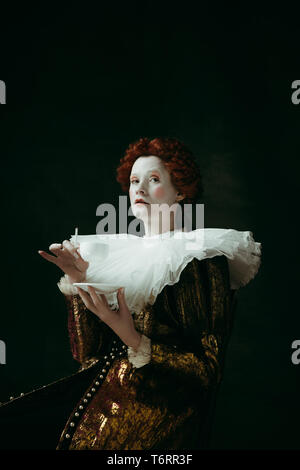 Medieval redhead giovane donna in oro di abbigliamento vintage come una duchessa tenendo una sigaretta e una tazza di caffè su sfondo verde scuro. Concetto di confronto delle epoche, la modernità e il rinascimento. Foto Stock