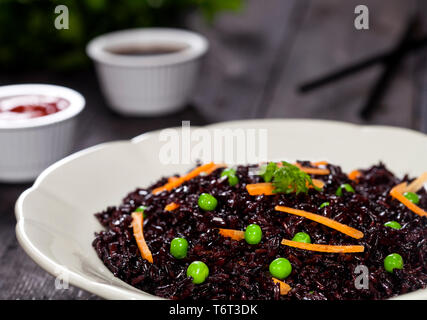 Cuocere il riso nero con verdure in una piastra con salse e bastoncini su uno sfondo di legno. Foto Stock