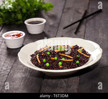 Cuocere il riso nero con verdure in una piastra con salse e bastoncini su uno sfondo di legno. Foto Stock