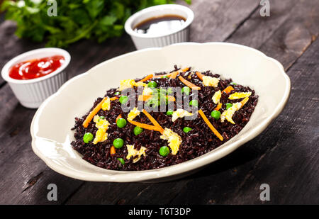 Cuocere il riso nero con verdure in una piastra con salse e bastoncini su uno sfondo di legno. Foto Stock