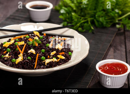 Cuocere il riso nero con verdure in una piastra con salse e bastoncini su uno sfondo di legno. Foto Stock
