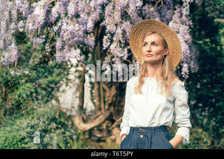 Bella donna europea bionda nel cappello di paglia contro lo sfondo di un albero con la fioritura del glicine Foto Stock