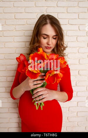 Giovane e bella ragazza al sesto mese di gravidanza in un luminoso abito rosso con un bouquet di tulipani sullo sfondo di un muro di mattoni. Foto Stock