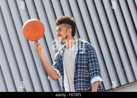 Giovane americano africano uomo in piedi isolato su una strada di città la filatura basket ball sul dito sorridente gioiosa vista laterale di close-up Foto Stock
