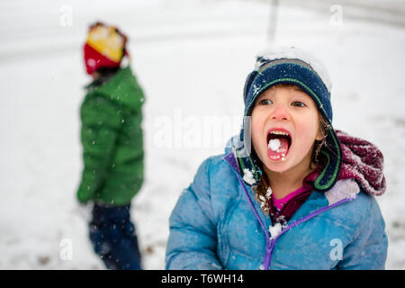 Una ragazzina in un fuzzy hat mangia la neve con il fratello in background Foto Stock
