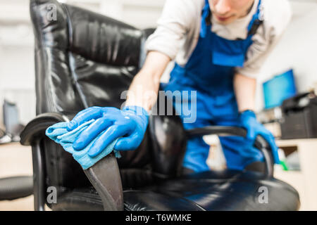 Giovane uomo in abbigliamento da lavoro e guanti di gomma pulisce la sedia da ufficio con attrezzature professionali. Foto Stock