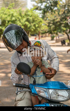 Sihanoukville, Cambogia - Marzo 15, 2019: Nero casco padre tiene toddler giovane figlio di fronte a lui sulla moto blu. Il verde delle foglie nel retro. Foto Stock