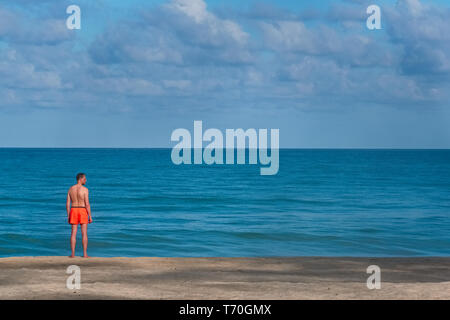 In un inizio di mattina a Bang Tao Beach, Phuket, Thailandia, un maschio solitario turista si affaccia sull'oceano Foto Stock