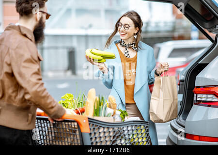 Giovane coppia elegante con carrello pieno di cibi freschi e prodotti di imballaggio in auto in un parcheggio all'aperto Foto Stock