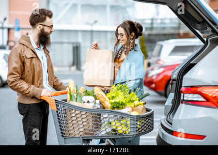 Giovane coppia elegante con carrello pieno di cibi freschi e prodotti di imballaggio in auto in un parcheggio all'aperto Foto Stock