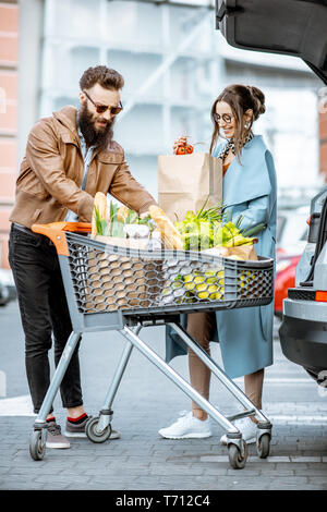 Giovane coppia elegante con carrello pieno di cibi freschi e prodotti di imballaggio in auto in un parcheggio all'aperto Foto Stock