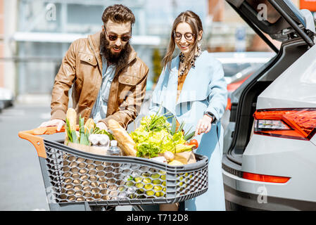 Giovane coppia elegante con carrello pieno di cibi freschi e prodotti di imballaggio in auto in un parcheggio all'aperto Foto Stock