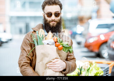 Felice l'uomo elegante con le borse della spesa piena di prodotti freschi e salutari all'aperto vicino al supermercato Foto Stock