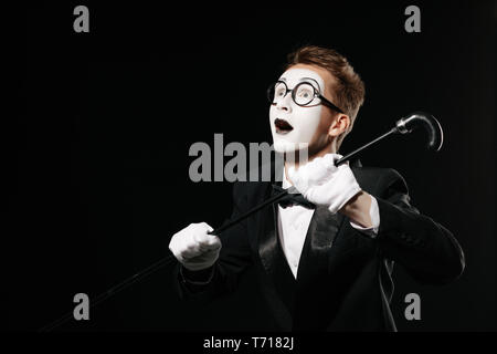 Sorpreso uomo mime in smoking e bicchieri in posa con bastone su sfondo nero Foto Stock