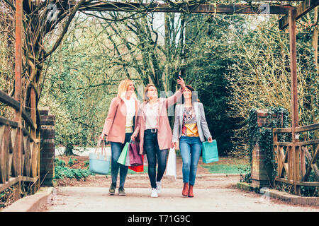 Tre giovani donne in un parco sorridente e prendersi cura delle borse della spesa che essi prendono un selfie mentre su a piedi - stare insieme, tempo libero, di amicizia e di consumo Foto Stock