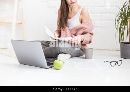 Una giovane donna felice si siede sul pavimento in un luminoso appartamento o di un ufficio interno e lavora su un portatile, freelancer ragazza al lavoro, il concetto di distanza Foto Stock