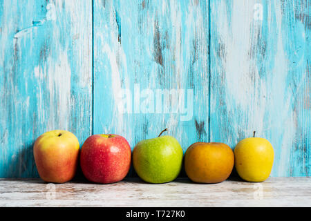 Alcune mele di diverse varietà, con colori diversi, mettere in linea contro un blu in legno rustico sfondo, con qualche spazio vuoto sulla parte superiore Foto Stock