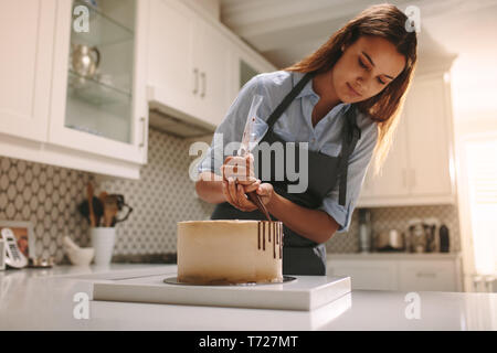 Lo chef femmina in grembiule decorare una torta in cucina. Chef pasticcere decorare la torta con la cioccolata liquida. Foto Stock