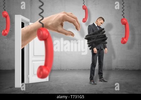 Triste imprenditore, legato con fili e un enorme mano che esce dalla porta dietro sempre pronti a catturare lui con quattro ricevitori telefono pendere dalla loro Foto Stock